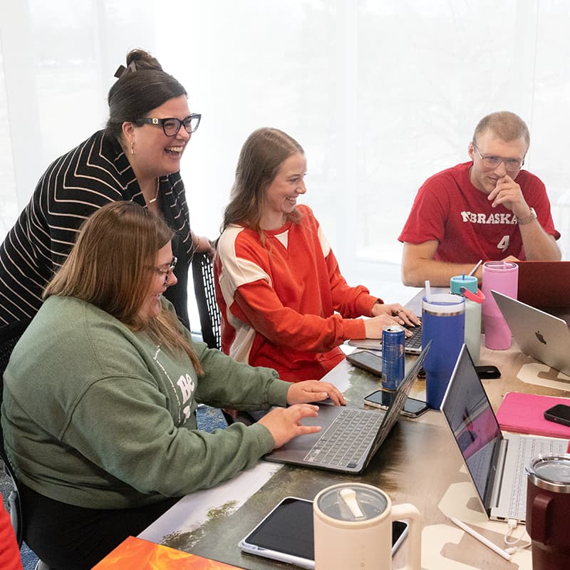 Group collaborating around laptops at a table in a bright room.