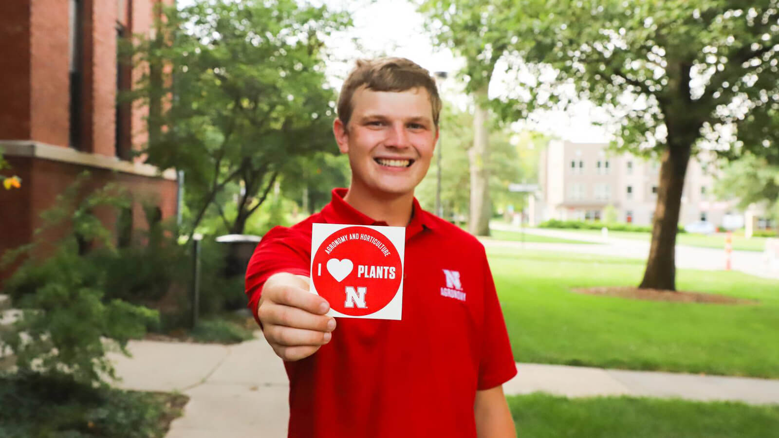 Man in red Agronomy polo shirt holds out a "I HEART Plants" sticker