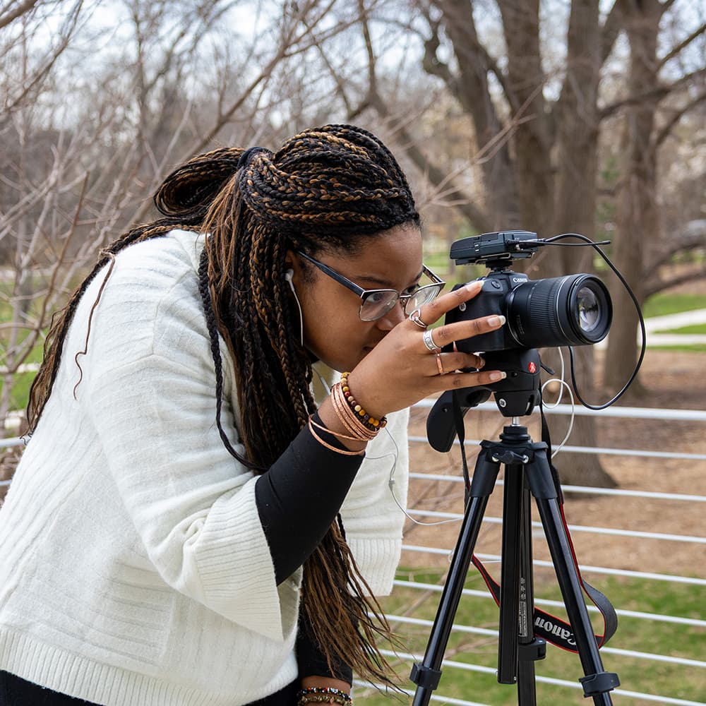 Photographer adjusting a camera on a tripod outdoors with bare trees in the background.