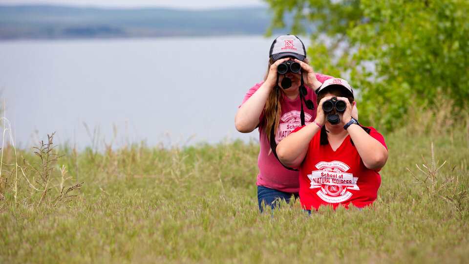 Two students standing in waist-high grass look through binoculars with a lake in the background.