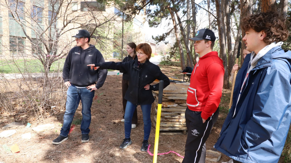 Students in Intro to Landscape Construction stand outdoors with Professor Kim Todd as she gives instructions for building a flagstone patio near some trees.