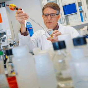Scientist in a lab coat using a pipette surrounded by bottles in a laboratory setting.