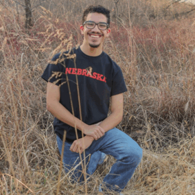 Person kneeling in dry grass wearing a black Nebraska shirt and blue jeans, surrounded by bare trees.