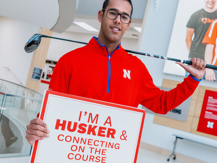Atirath Deb roy stands in Howard L. Hawks Hall holding a golf club and a sign that reads, "I'm a Husker & connecting on the course." 