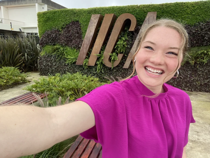 Lexi Bodlak takes a selfie in front of a wall of greenery featuring the IICA logo
