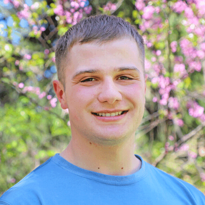 Man in a blue shirt standing outdoors with blooming pink flowers in the background.