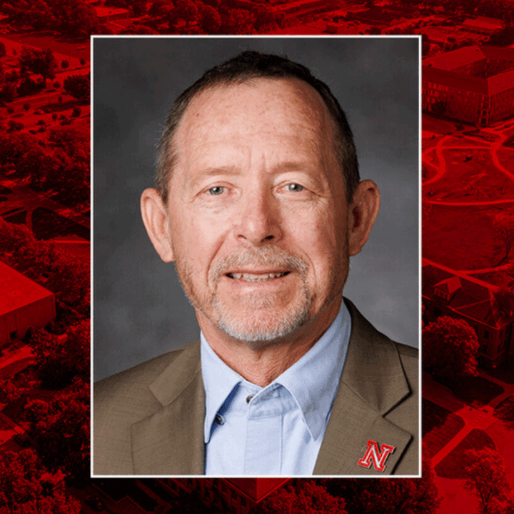 Man with short hair and beard wearing a beige blazer and light blue shirt, with a red "N" pin.