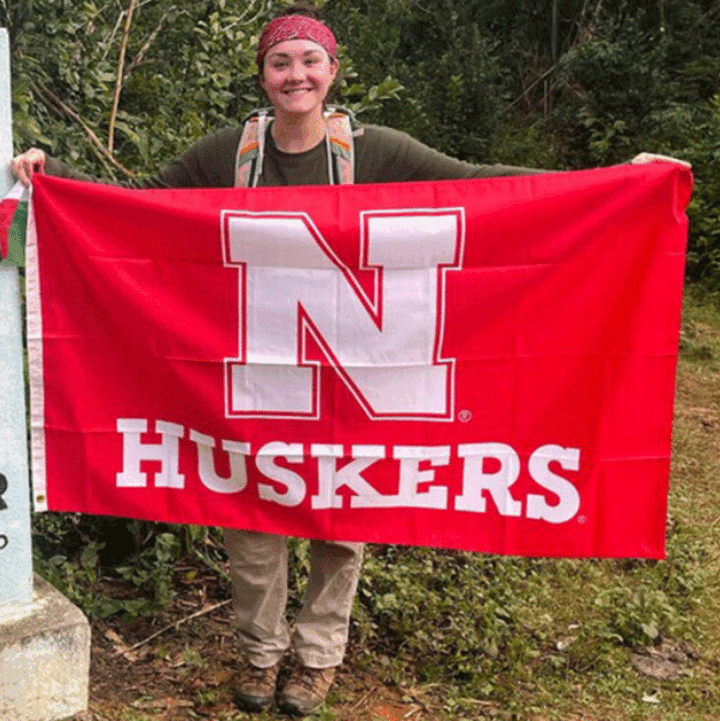 Person outdoors holding a red Nebraska Huskers flag, smiling with greenery in the background.